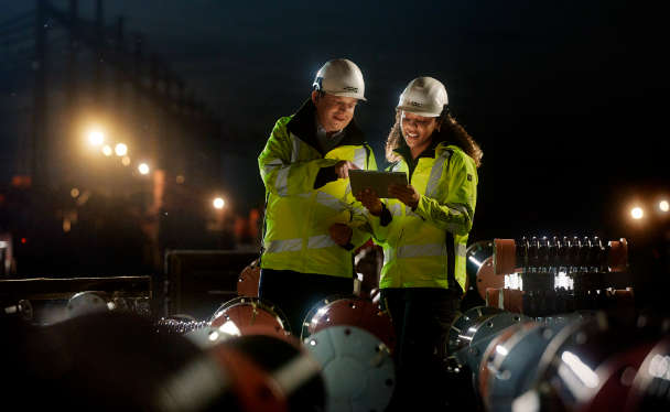 Ein Mitarbeiter und eine Mitarbeiterin stehen in der Dämmerung im Umspannwerk zwischen Ersatzteilen und blicken auf ein Ipad.