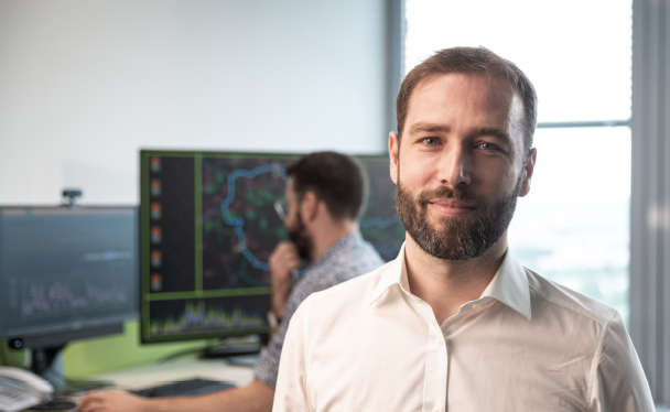 Ein Mann in weißem Hemd blickt in die Kamera, hinter ihm ein weiterer Mann, der an einem Schreibtisch sitzt und auf einen Bildschirm blickt.