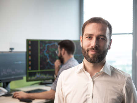 Ein Mann in weißem Hemd blickt in die Kamera, hinter ihm ein weiterer Mann, der an einem Schreibtisch sitzt und auf einen Bildschirm blickt.