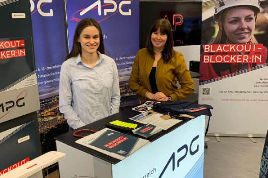 Zwei APG-Mitarbeiterinnen hinter einem Pult eines APG-Messestandes. Sie blicken lächelnd in die Kamera.