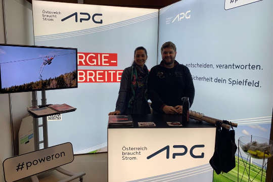 Two APG employees behind a desk at an APG trade fair stand. They smile at the camera.