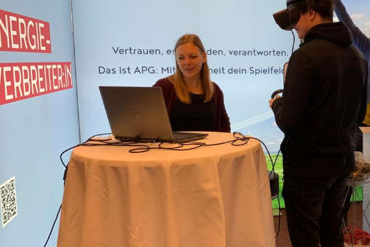 Eine APG-Mitarbeiterin hinter einem Tisch eines APG-Messestandes. Sie unterhält sich mit einem schwarz gekleideten Mann mit Kappe und blickt dabei auf ein Notebook.