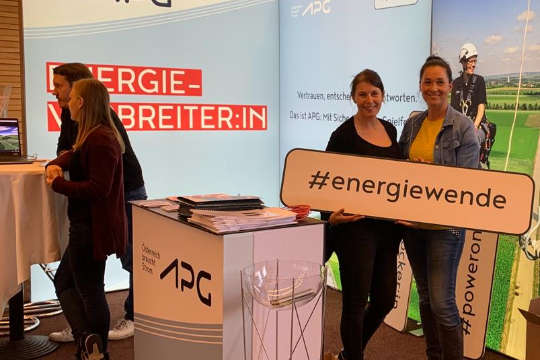 Vier APG-Mitarbeiterinnen auf einem APG-Messestand. Zwei stehen seitlich zur Kamera an einem Tisch angelehnt, zwei halten in die Kamera lächelnd ein Schild mit der Aufschrift "#energiewende"