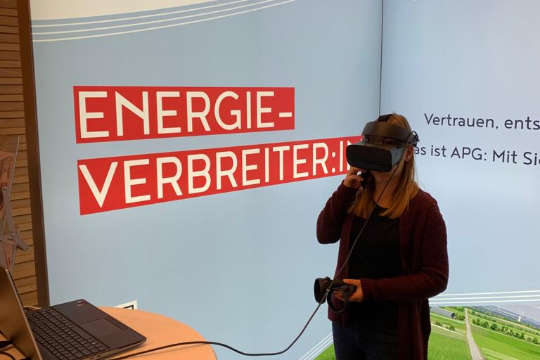 Eine APG-Mitarbeiterin vor einem weißen Tisch auf einem APG-Messestand. Sie trägt eine VR-Brille am Kopf und hält einen Joystick in der Hand.