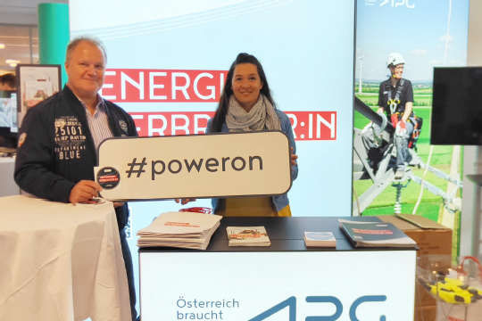 Zwei APG-Mitarbeiter stehen vor einem Pult bei einem APG-Messestand und halten in die Kamera lächelnd, gemeinsam ein Schild mit der Aufschrift "#poweron".