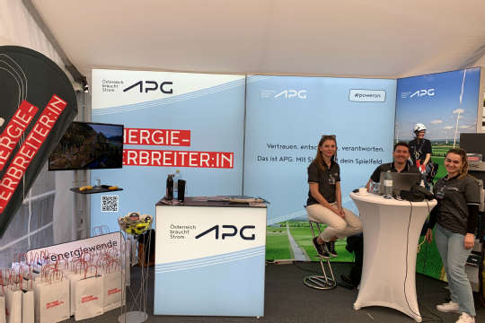 Three APG employees at an APG exhibition stand. One is sitting on a bar stool, two others are standing at a bar table with camera equipment in their hands. They are all smiling at the camera.