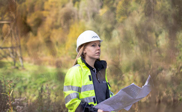 Eine APG-Mitarbeiterin Sicherheitskleidung steht vor einem Strommasten, der in eine Waldlandschaft eingebettet ist und hält dabei ein großes Blatt Papier in den Händen.